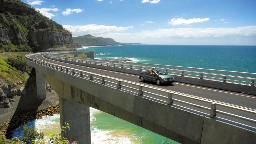 Long view of car driving along Sea Cliff Bridge, Grand Pacific Drive, with coastal views in background, Illawarra