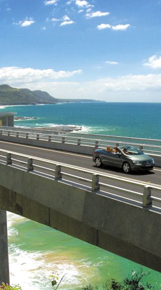 Long view of car driving along Sea Cliff Bridge, Grand Pacific Drive, with coastal views in background, Illawarra