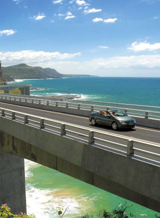 Long view of car driving along Sea Cliff Bridge, Grand Pacific Drive, with coastal views in background, Illawarra