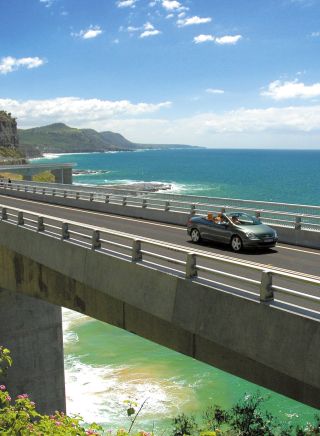 Long view of car driving along Sea Cliff Bridge, Grand Pacific Drive, with coastal views in background, Illawarra